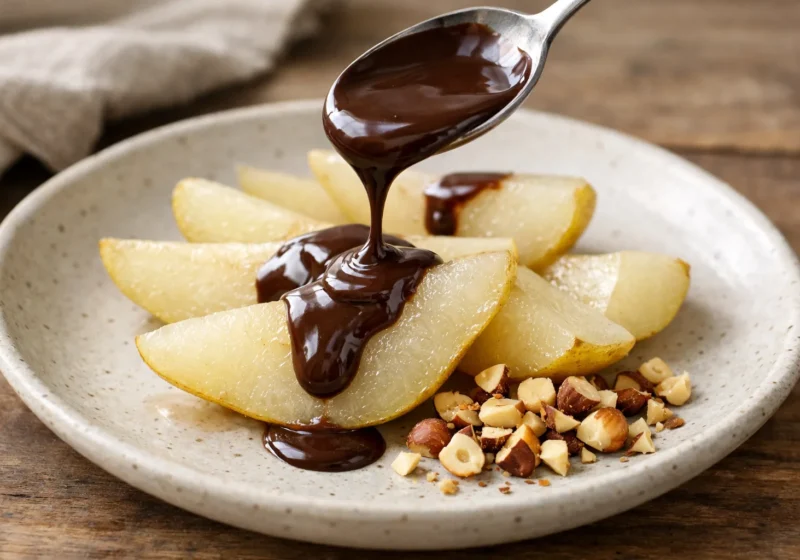 Une cuillère verse une sauce au chocolat sur des quartiers de poire, avec des éclats de noisette dans l’assiette.
